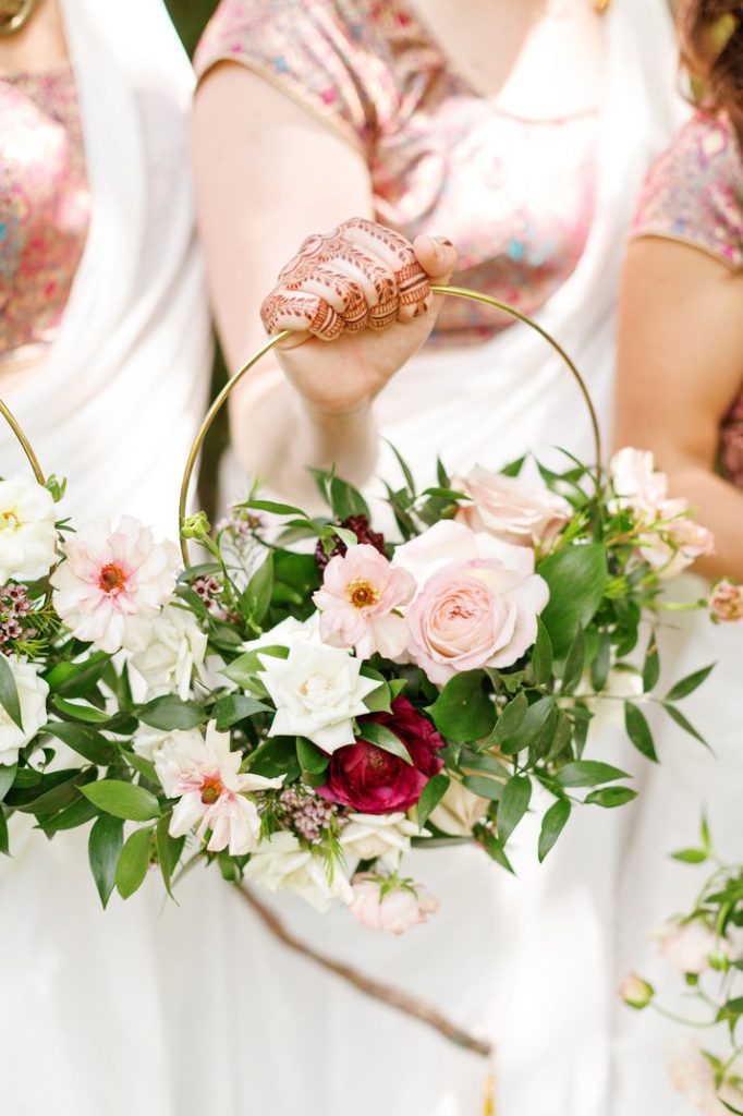 In questa foto una sposa tiene nella mano destra un bouquet a cerchio decorato con rose rosa, bianche e rosse, foglie di vischio 