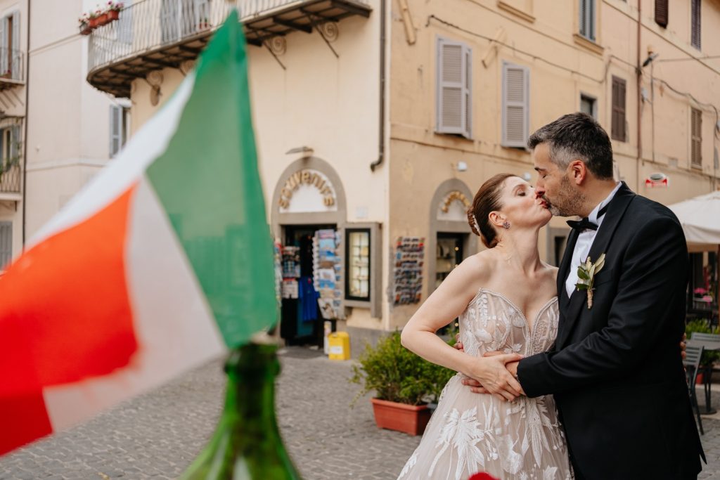 In questa foto una coppia di sposi si bacia davanti a un negozio di souvenir, con sullo sfondo una bandierina dell'Italia: lo scatto è stato realizzato da Sara Sganga, tra i migliori fotografi di matrimonio a Roma