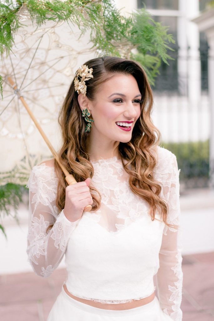 In questa foto una sposa sorride tenendo un ombrellino pizzo sotto un albero. Indossa un abito da sposa con crop top e maniche lunghe in pizzo rebrodé. Ha i capelli sciolti e mossi fermati su un lato da un accessorio colore oro 3D. Indossa orecchini pendenti verde smeraldo