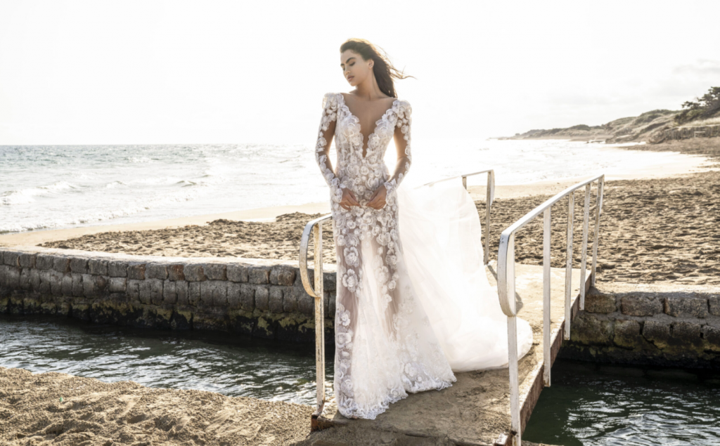 In questa foto una modella vicino al mare indossa un abito da sposa Alessandro Angelozzi 2021 con profondo scollo a V e ricami in pizzo floreale