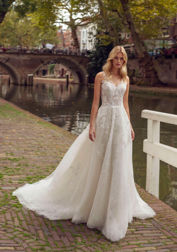 In questa foto una modella passeggia sulla riva di un fiume indossando un abito da sposa Modeca 2021