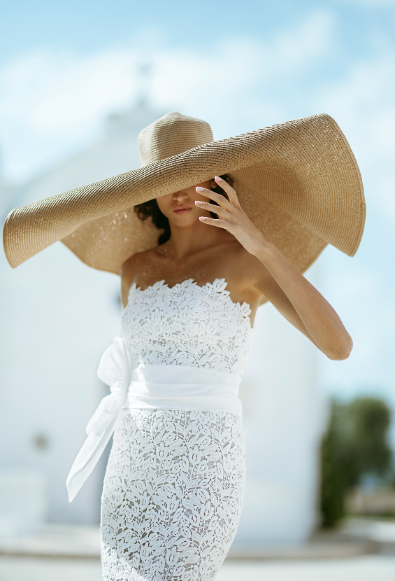 In questa foto una modella indossa un cappello in paglia dal diametro circolare grandissimo e un abito da sposa in pizzo di Luisa Sposa 2020/2021