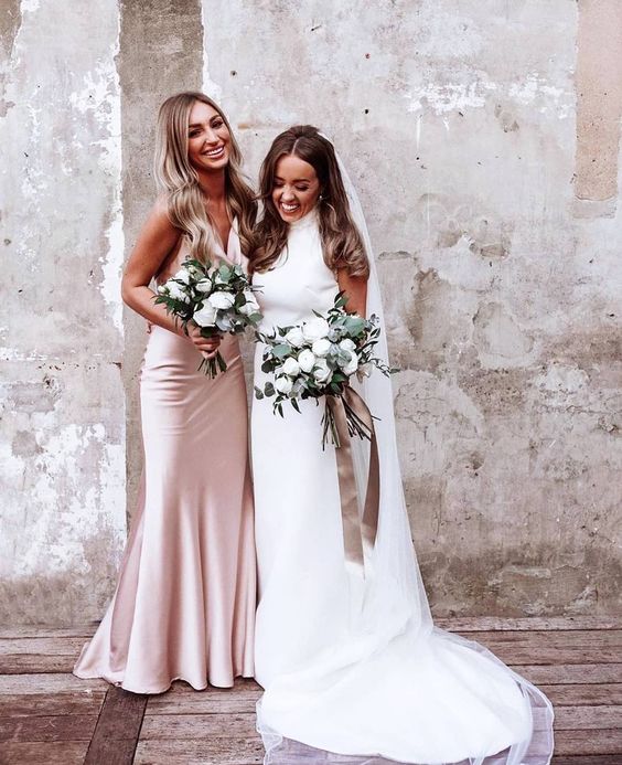 In questa foto una sposa con la sua damigella d'onore vestita con un abito sottoveste in seta rosa, sorridenti e con in mano i loro bouquet floreali 