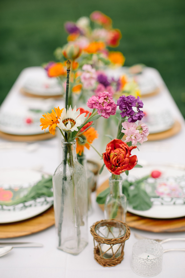 In questa foto composizioni floreali per i centrotavola di matrimonio allestito sul prato realizzati con bottiglie di vetro e fiori di campo colorati