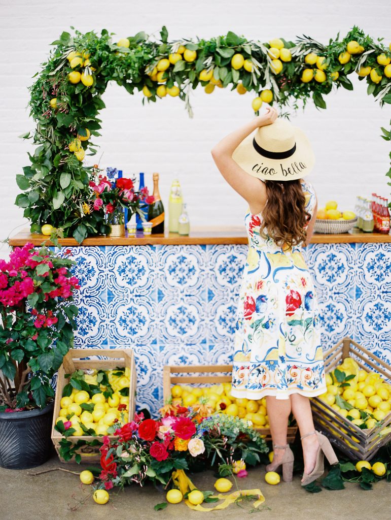 In questa foto una modella con un vestito senza maniche in stile siciliano si avvicina ad un angolo di bevande. Il banchetto è decorato con maioliche blu, un arco e cassette piene di limoni 