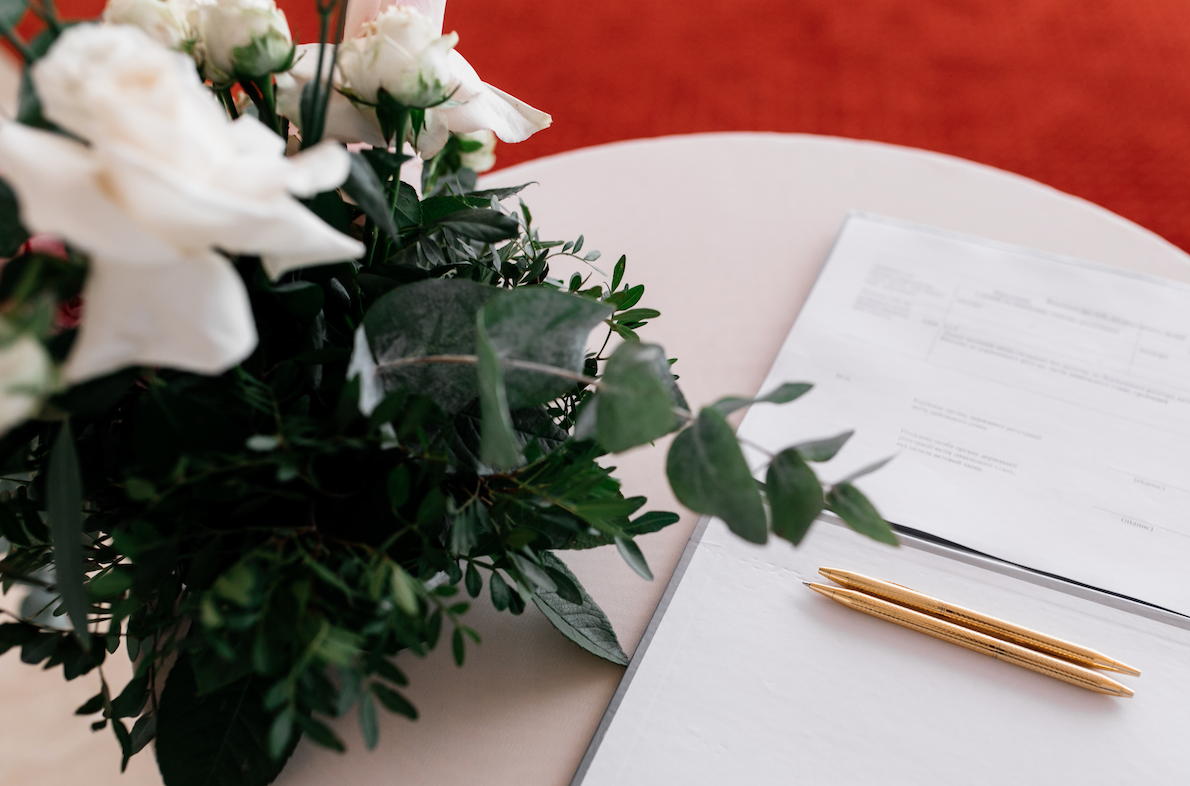 In questa foto un bouquet di nozze, due penne e un documento di matrimonio sopra un tavolino