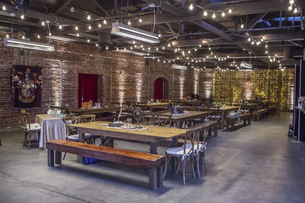 In questa foto una sala in stile industriale con tavoli imperiali in legno e panche in coordinato perfetti per riprodurre la Sala Grande di Hogwarts per un matrimonio a tema Harry Potter. Sulle pareti sono affissi gli stendardi delle case.