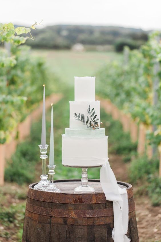 In questa foto una torta di matrimonio bianca e verde salvia su una botte in legno, in mezzo ad un vigneto