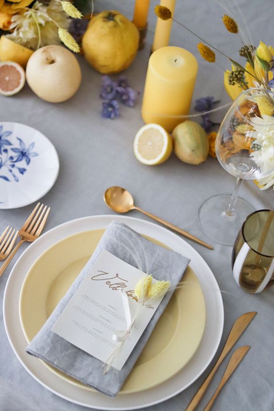 In questa foto una mise en place vista dall'alto per un matrimonio a tema giallo e grigio con posate in oro, candele gialle e limoni come centrotavola. II sottopiatto è bianco mentre il piatto è in giallo tenue e il tovagliolo in grigio chiaro a contrasto