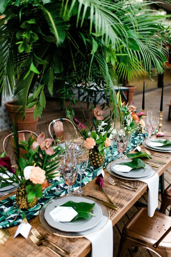 In questa foto un tavolo matrimonio imperiale in legno visto dall'alto in stile jungle con con palme, orchidee e ananas come centrotavola. I piatti sono bianchi, i sottopiatti trasparenti con bordi colore oro e ci sono foglie larghe come segnaposto 