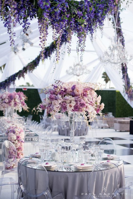 In questa foto un allestimento di nozze con tovagliato color lavanda e centrotavola in coordinato con cristalli e fiori bianco. Il ricevimento si svolge sotto un tendone di stoffa trasparente con archi di glicine e lampadari in cristallo