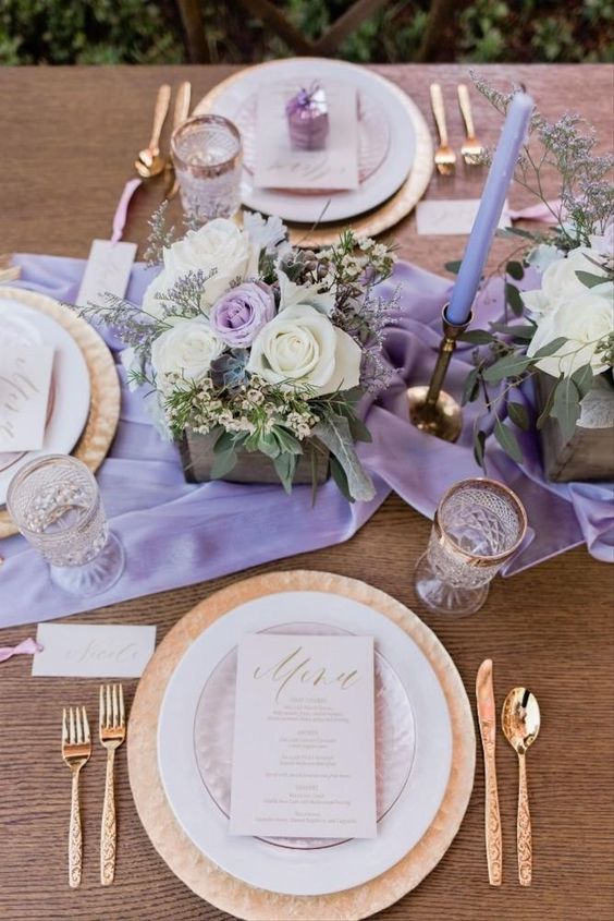 In questa foto una mise en place boho chic e romantica con runner lilla, centrotavola di rose bianche, piatti e posate in oro