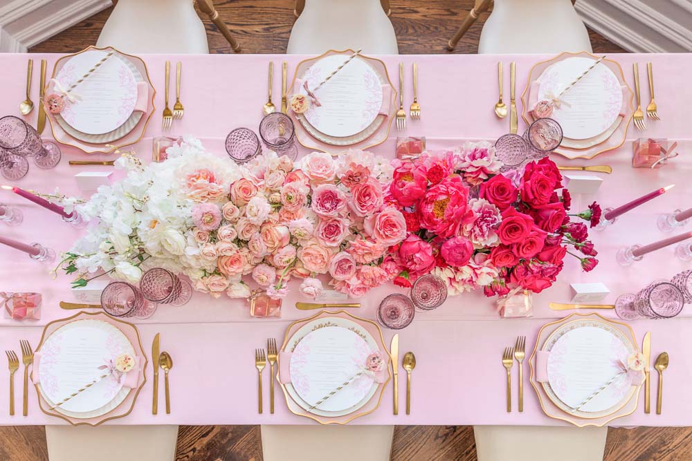 In questa foto un runner floreale visto dall'alto utilizzato come centrotavola per un matrimonio. I fiori utilizzati sono disposti in gradazione di colore dal bianco al rosa al fuxia