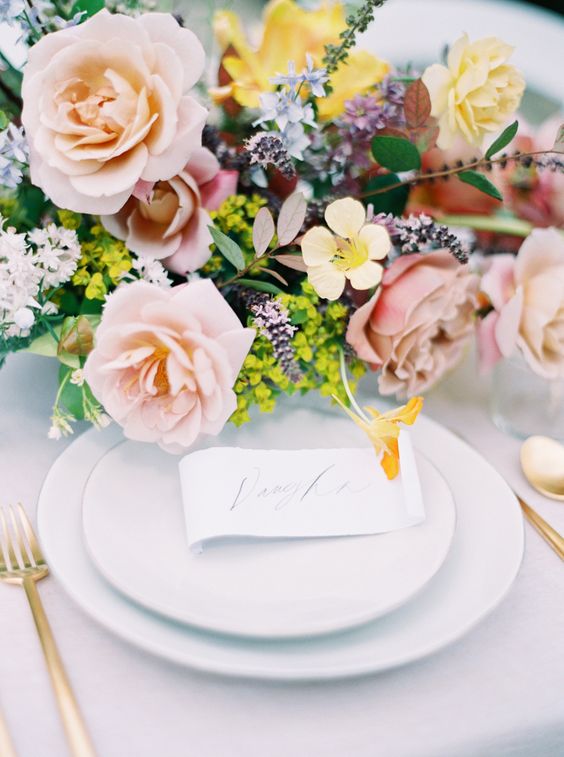 In questa foto una mise en place elegante con tovagliato e piatti bianchi, posate oro e centrotavola di rose color pesca e fiori di campo