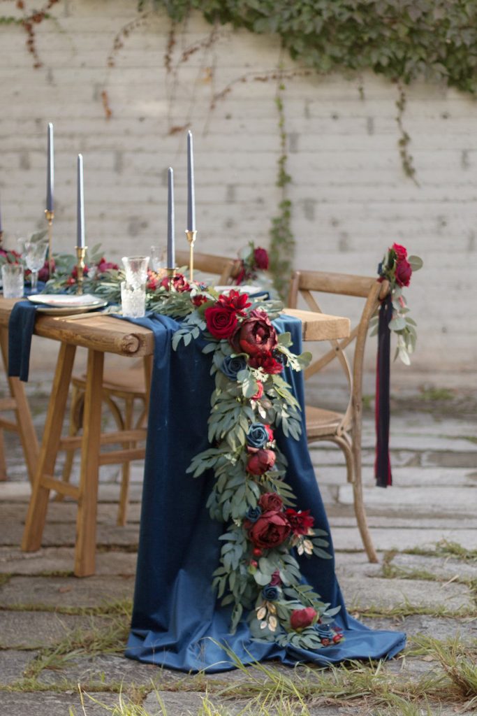 In questa foto l'allestimento di un tavolo matrimonio in legno nudo con runner di stoffa blu e runnere di foglie e grandi fiori blu e rossi