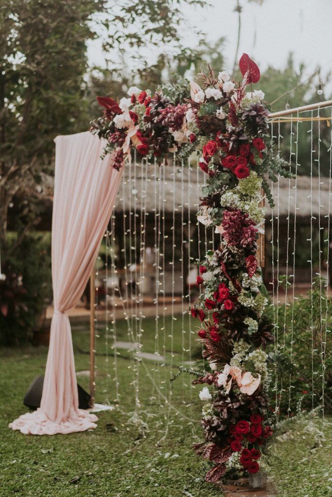 In questa foto un arco per un rito nuziale su un prato di fiori rossi, bianchi, verdi e rosa con tendaggi rosa confetto e e catenelle di cristalli