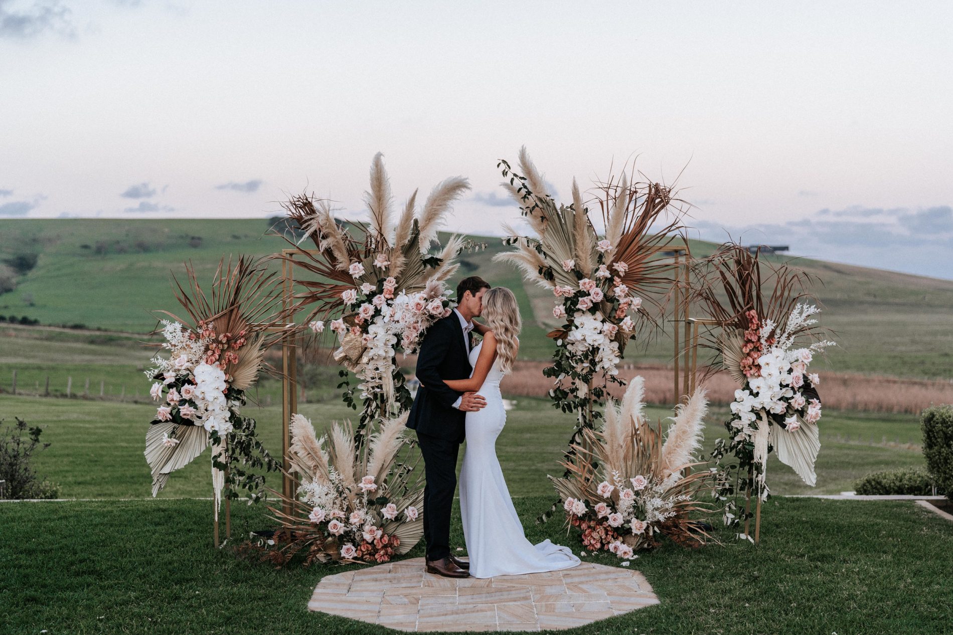 In questa foto due sposi che si baciano dopo la celebrazione del loro matrimonio in stile boho chic. Alle spalle, al posto dell'altare, un arco simbolico con pampas e rose