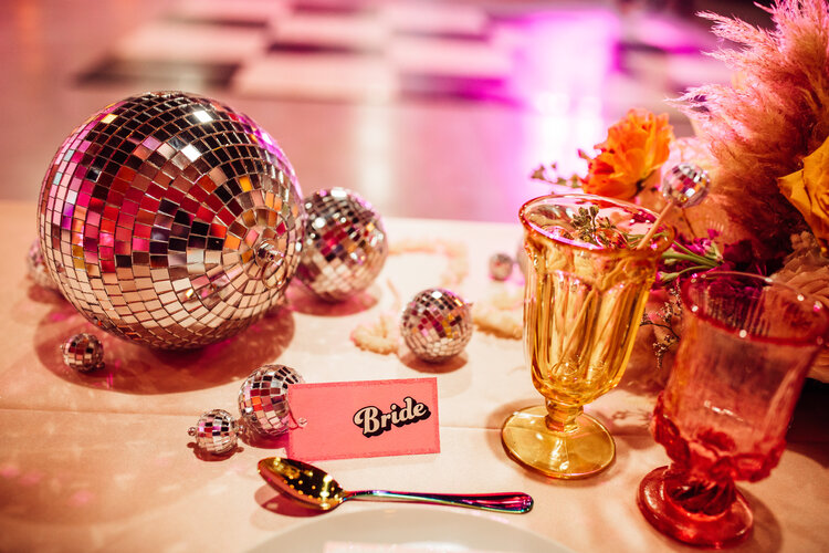 In questa foto il tavolo degli sposi con una palla stroboscopica colore argento, la targhetta rosa della sposa e bicchieri colorati. Un allestimento ideal per un matrimonio a tema anni 80