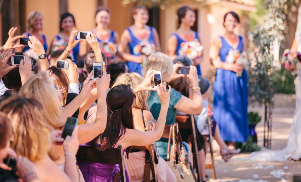 In questa foto gli invitati di un matrimonio scattano con i loro smartphone foto agli sposi durante la cerimonia di nozze. Sullo sfondo si intravedono fuori fuoco le damigelle d'onore vestite di blu con bouquet arancioni