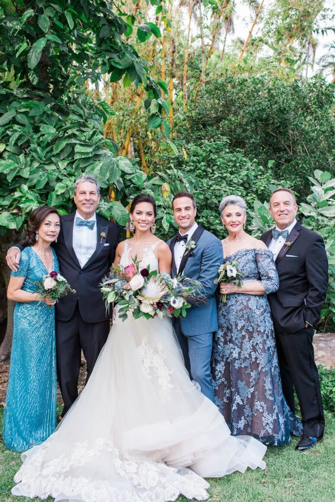 In questa foto due sposi posano in un giardino con i rispettivi genitori tutti con abiti e dettagli in blu. La sposa indossa un abito ampio e tiene tra le mani un bouquet a braccio di fiori colorati