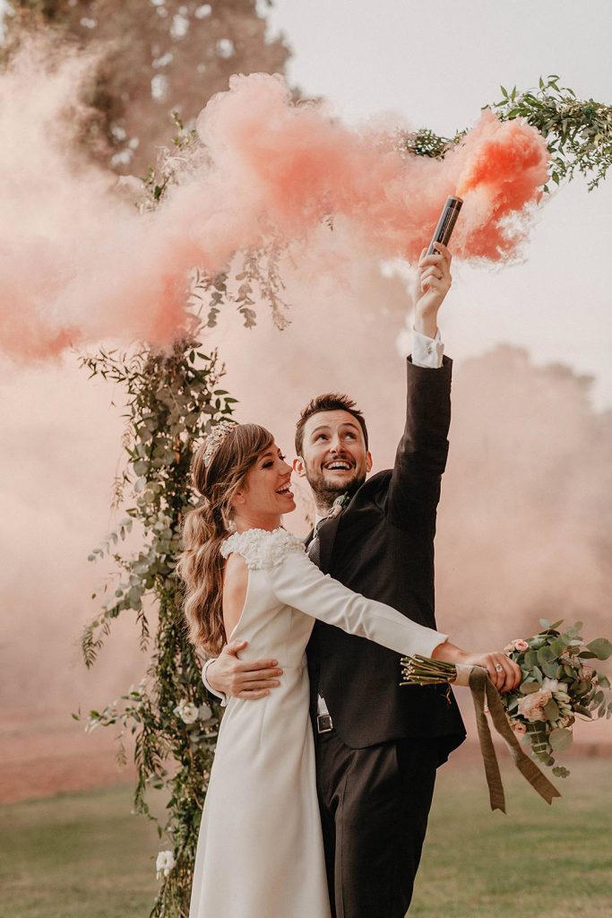 In questa foto due sposi sorridenti si abbracciano su un prato mentre lo sposo agita un bengala di fumo rosso