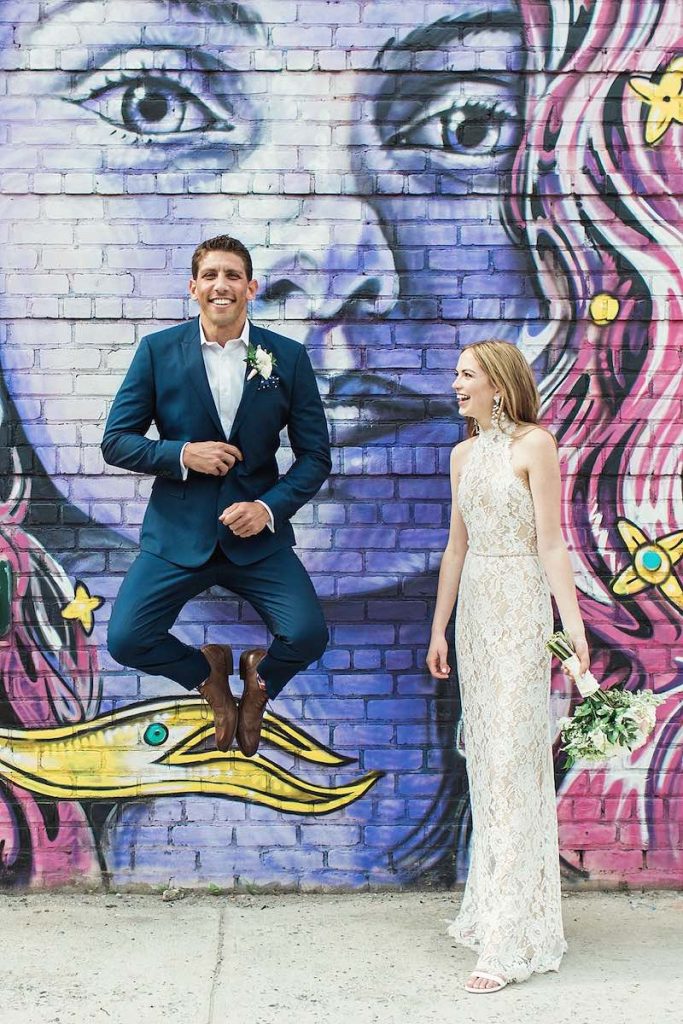 In questa foto matrimonio due sposi davanti ad un murales. Lo sposo salta in aria con le gambe piegate e sorride. la sposa lo guarda e ride