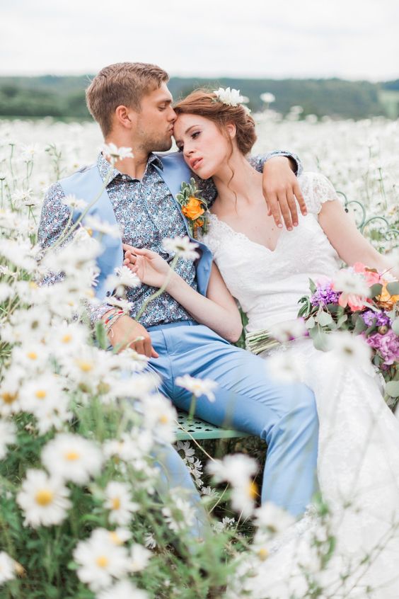 In questa foto due sposi posano dopo la cerimonia immersi in un prato di margherite. La sposa ha la testa appoggiata sulla spalla dello sposo, vestito d'azzurro, che la bacia sulla fronte