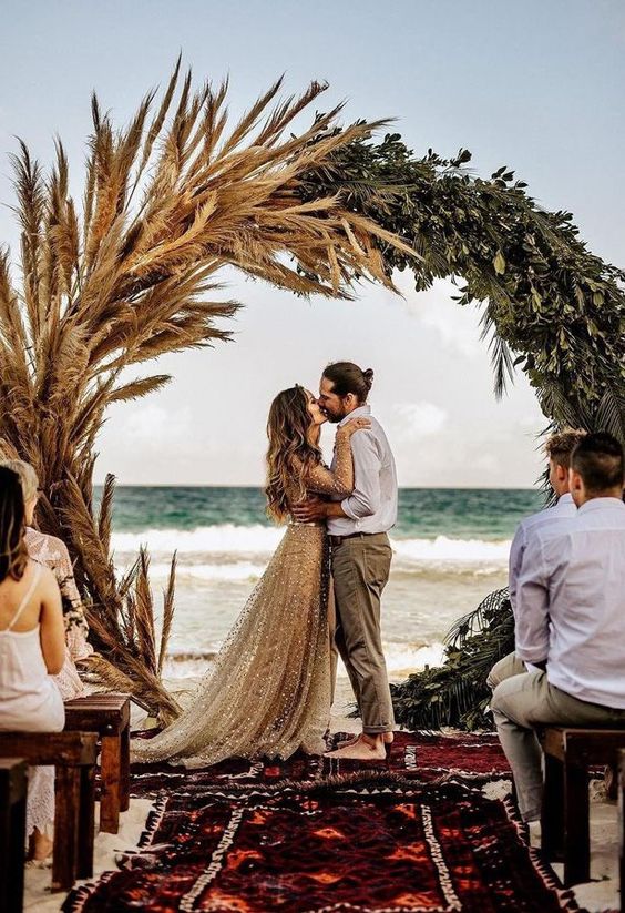 In questa foto due sposi si baciano alla fine della cerimonia di matrimonio sulla spiaggia in riva al mare davanti ad un arco di pampas e foglie mentre tutti gli ospiti li guardano