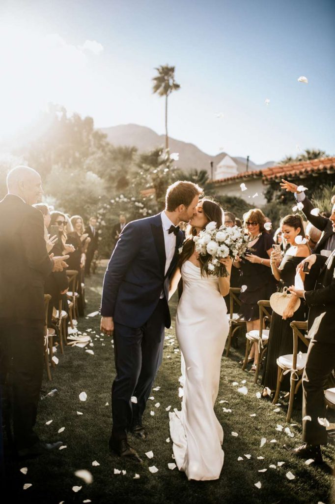 In questa foto due sposi si baciano mentre camminano tra gli invitati in piedi lungo il corridoio della location sul prato dove è stata celebrata la cerimonia civile. La sposa indossa un abito a sirena effetto lingerie e tiene in mano un bouquet di peonie bianche