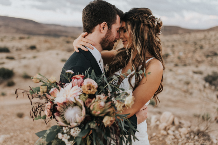 In questa foto due sposi posano abbracciati in una foto di matrimonio realizzata in un campo di grano. La sposa ha i capelli sciolti, tiene una mano appoggiata dietro la nuca del suo sposo e nell'altra mano tiene un bouquet a braccio di fiori di campo