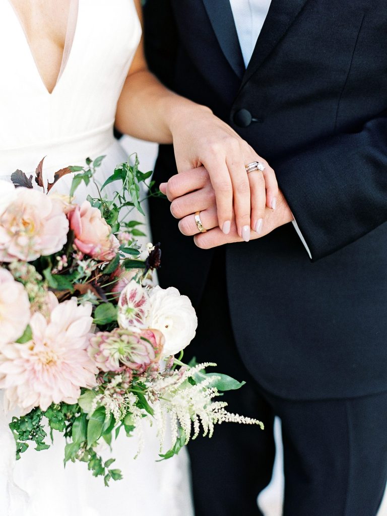 In questa il dettaglio delle mani strette di due sposi. L'attenzione si concentra sulle fedi e il bouquet di fiori rosa cipria della sposa