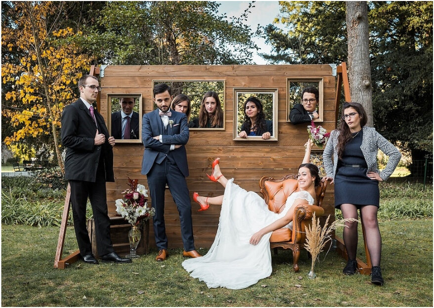 In questa foto due sposi in una posa divertente insieme agli amici davanti ad un photo booth creato con uno steccato di legno e delle cornici su un prato. La sposa è seduta su una poltrona con le gambe e il bouquet alzati mentre lo sposo è in piedi. Accanto a loro due amici. Dalle cornici sbucano altri 5 amici