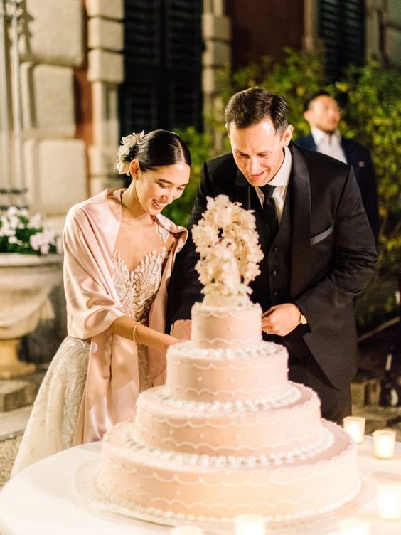 In questa foto due sposi sorridenti tagliano la loro torta nuziale a quattro piani, rosa e con un cake topper floreale ripresa in primo piano. La sposa indossa uno scialle in coordinato e capelli raccolti