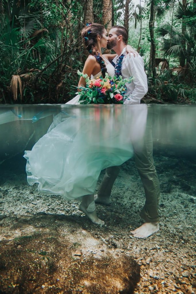 In questa foto due sposi posano mentre si baciano abbracciati dentro un lago immersi in acqua fino al busto. La sposa ha i capelli intrecciati con petali blu e tiene tra l mani un bouquet di foglie e fiori rosa, gialli e blu