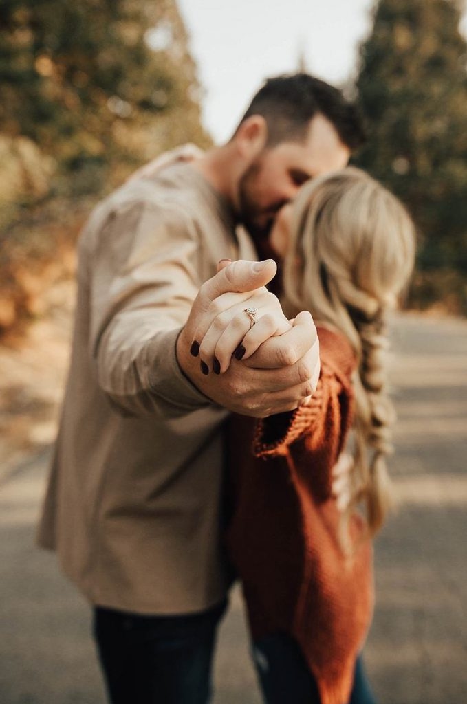 In questa foto due fidanzati, ritratti fuori fuoco, si baciano mentre abbracciati si tengono per mano con le braccia tese verso l'obiettivo mostrando in primo piano l'anello di fidanzamento