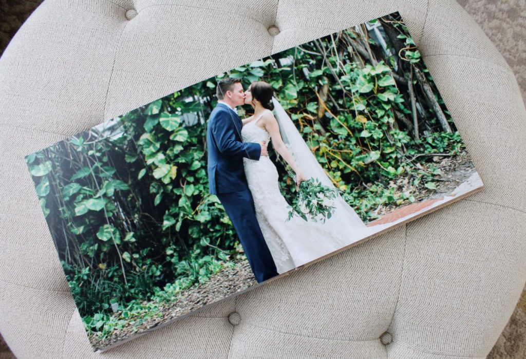 In questa foto un fotoalbum di matrimonio aperto sulla foto di due sposi che si baciano in un bosco e poggiato su un pouf beige 