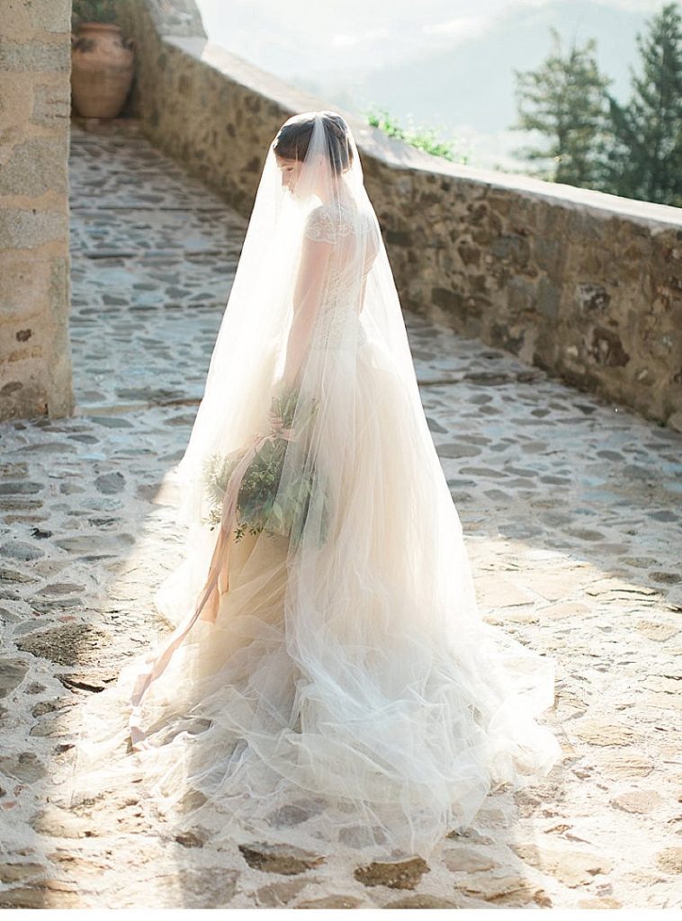 In questa foto una sposa viene ritratta sotto il suo velo mentre, filtrando la luce, si intravedono il bouquet legato da un nastro rosa, la sua acconciatura e l'ampia gonna in tulle