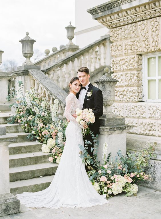 In questa foto due sposi abbracciati posano davanti ad una scalinata guardando verso il fotografo. Lei tiene in una mano il bouquet di fiori bianchi e rosa. La scalinata è decorata con fiori bianchi e rosa e foglie verdi