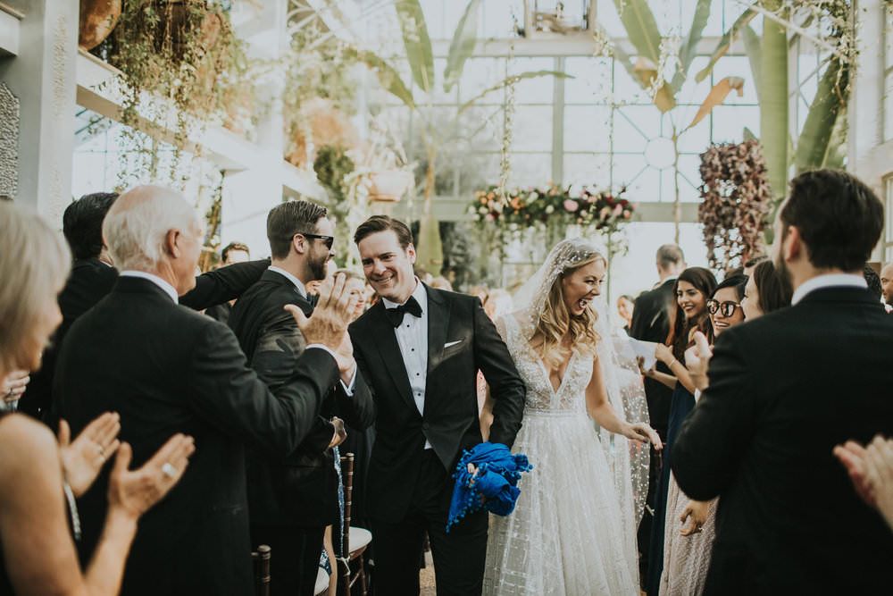 In questa foto due sposi lungo la navata di una chiesa decorata con fiori escono dopo la cerimonia del loro matrimonio salutando, sorridenti, amici e parenti