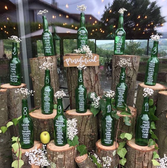 In questa foto un tableau mariage realizzato con bottiglie di birra verdi vuote su cui sono riportate le liste degli ospiti divisi per tavolo. Le bottiglie poggiate su tronchi d'albero decorati con fiori, edera e mele sono perfette per un matrimonio a tema vino