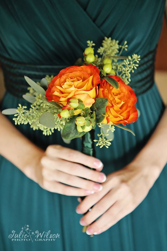 In questa foto una damigella indossa un abito verde scuro e tiene tra le mani un piccolo bouquet di rose arancioni, bacche verdi e piccole foglioline