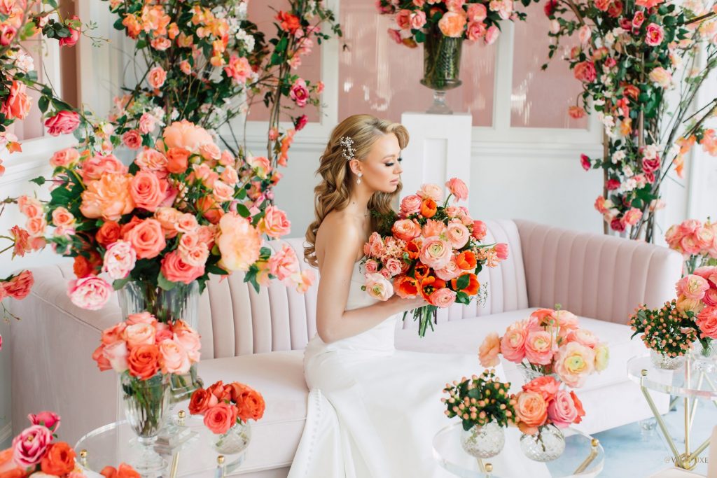 In questa foto una modella vestita da sposa posa per uno shooting fotografico a tema corallo seduta su un divano rosa e circondata da rose in tutta la stanza in coordinato.  Guarda il bouquet di rose, papaveri e ranuncoli che stringe tra le mani