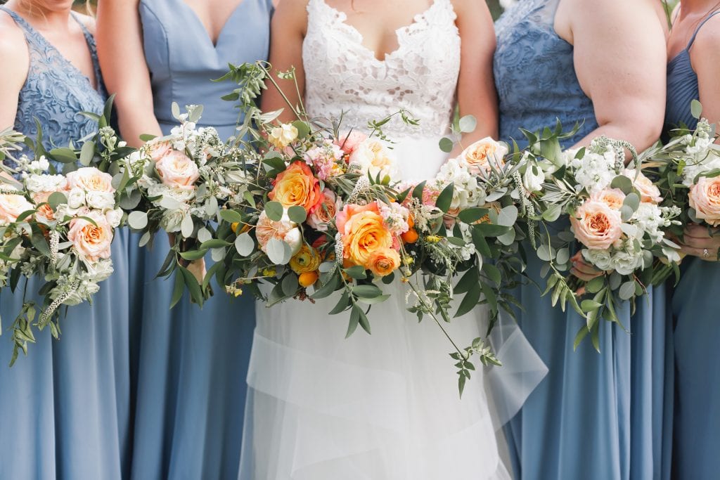 In questa foto la sposa con le sue damigelle riprese dal collo al busto tengono in mano i loro bouquet con rose arancioni e colore pesca e rametti verdi. Le damigelle indossano abiti colore azzurro polvere