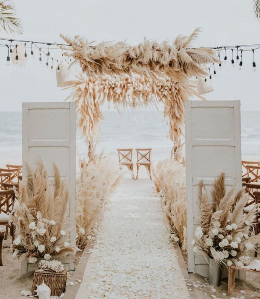 In questa foto un allestimento di un rito nuziale sulla spiaggia con pampas e fiori bianchi e avorio. Sullo sfondo sono disposte due sedie per gli sposi