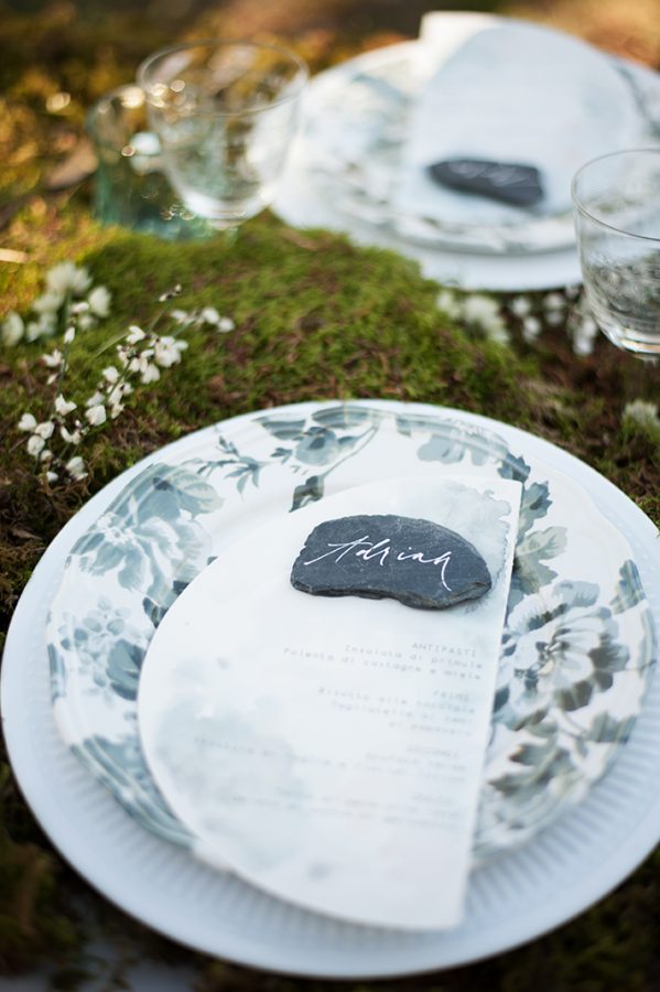 In questa foto una mise en place nei colori del verde scuso e del bianco perfetta per un matrimonio in un bosco. Al posto del tovagliato è usato del muschio decorato con fiori bianchi, sassolini piatti neri sono usati come segnaposto su cui è riportato il nome di ogni ospite. Ogni segnaposto è appoggiato sul menù stampato su carta trasparente a forma di mezza Luna e su piatti di porcellana bianca decorata con fiori verdi