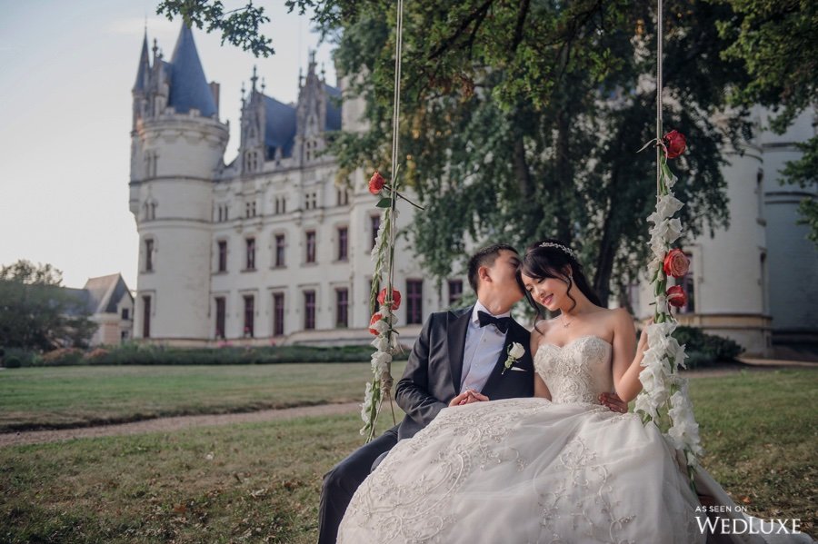 In questa foto lo sposo bacia sulla testa la sposa mentre sono seduti su un'altalena decorata con fiori bianchi e arancioni. Sullo sfondo si vede un prato e un castello