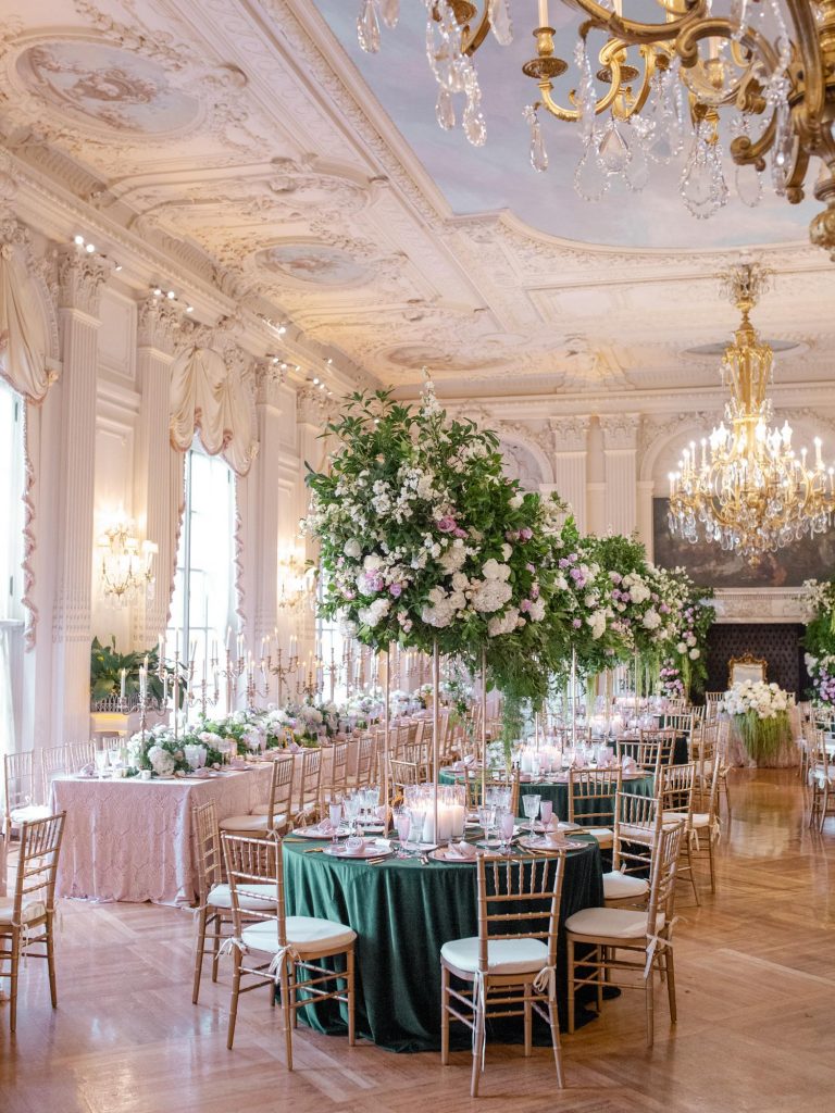 In questa foto un matrimonio allestito nella sala di una villa. Il tovagliato è verde bosco e rossa mentre la mise en place oro. Come centrotavola sono usate grandi composizioni floreali in coordinato appoggiate a lunghi piedistalli dorati di forma prismatica