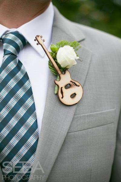 In questa foto una bottoniera su un abito grigio con una rosa bianca, rametti verdi e una piccola chitarra in legno intagliato