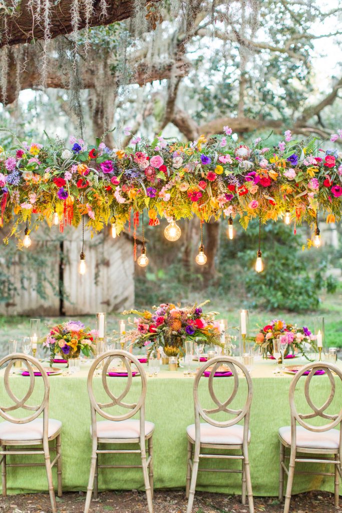 In questa foto un allestimento per un matrimonio in primavera con sedie in legno stile Shabby Chic, tovagliato colore verde pistacchio e fuxia. Come centrotavola sono usati cesti di fiori arancioni, rosa e viola. Gli stessi fiori sormontano tutto il tavolo con un arco orizzontale a cui sono legate lampadine che scendono in verticale