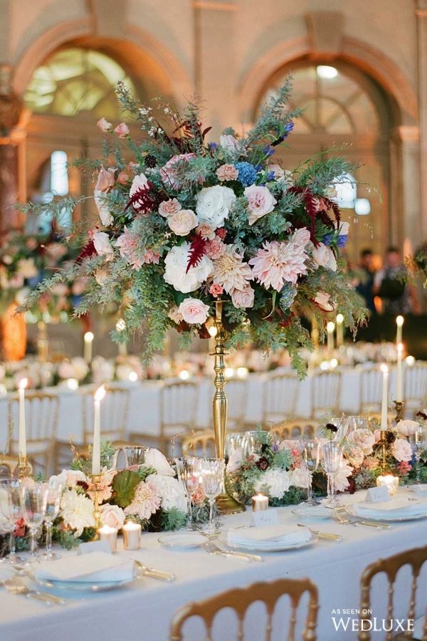 In questa foto un allestimento di un matrimonio in una sala elegante. Il tovagliato e le porcellane sono bianche, le posate in argento, le sedie colore oro. Sulla tavola imperiale è disposto un runner di fiori bianchi, bordeaux e rosa cipria. Presenti anche candele bianche accese centrotavola alti con composizioni di fiori in coordinato e felce